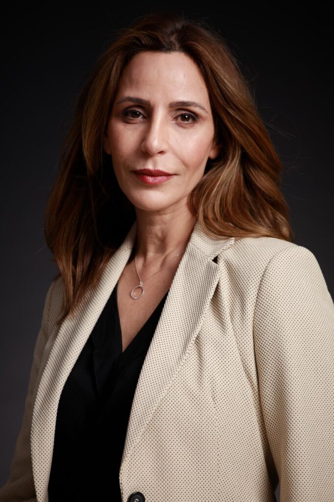 Real estate properties in caesarea Real estate properties in caesarea Woman with long brown hair and layered necklaces wearing a black top, smiling and leaning forward against a dark background.
